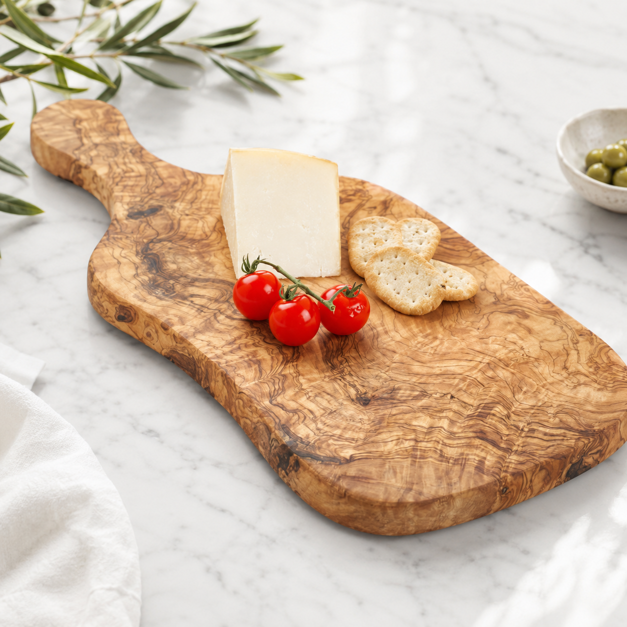 Handgefertigtes Servierbrett aus Olivenholz mit natürlicher Maserung, belegt mit Käse, Cherrytomaten und Crackern auf hellem Marmortisch.