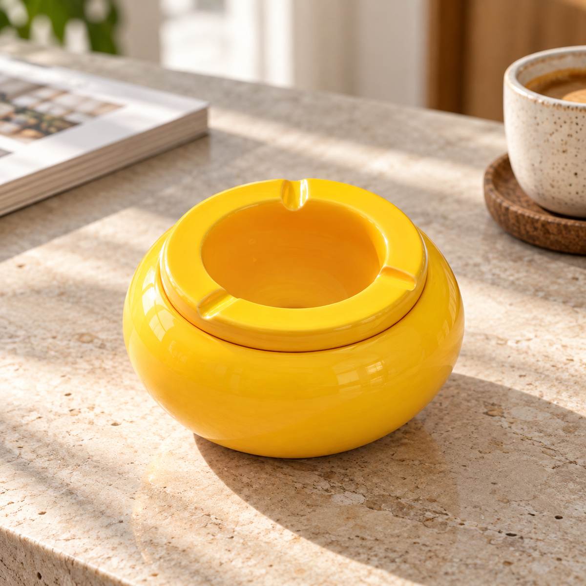 Leuchtend gelber Keramik-Aschenbecher mit glänzender Oberfläche auf einem sonnigen Steintisch neben Kaffeetasse und Wohnmagazin.