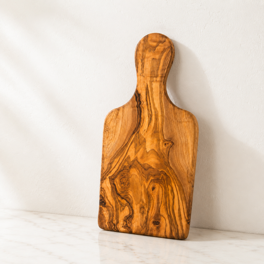 Handgefertigtes Schneidebrett aus massivem Olivenholz mit natürlicher Maserung vor heller Wand – MediterraneanArt Schweiz